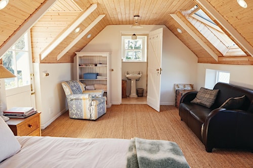 Chambre mansardée avec plafond en bois, lit, fauteuil, canapé et salle de bain attenante