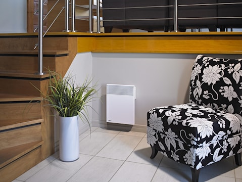 Chauffage électrique mural blanc installé dans un salon moderne, à côté d’un fauteuil à motif floral et d’une plante décorative