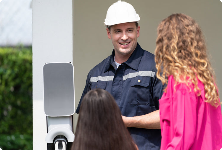 Technicien en uniforme souriant à deux clientes devant une borne de recharge blanche installée sur un mur extérieur résidentiel.