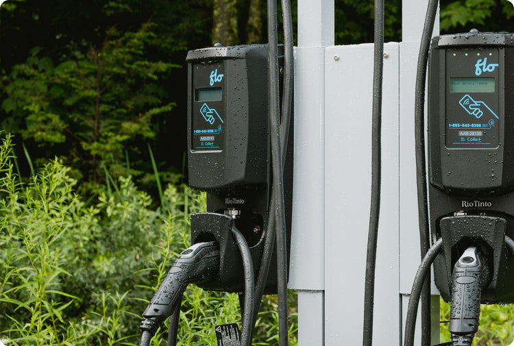Deux bornes de recharge FLO installées sur un poteau blanc extérieur, câbles de recharge suspendus, couvertes de gouttes de pluie.